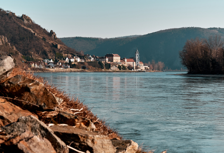 Malerische Flusslandschaft mit historischem Gebäude und bewaldeten Hügeln im Hintergrund.