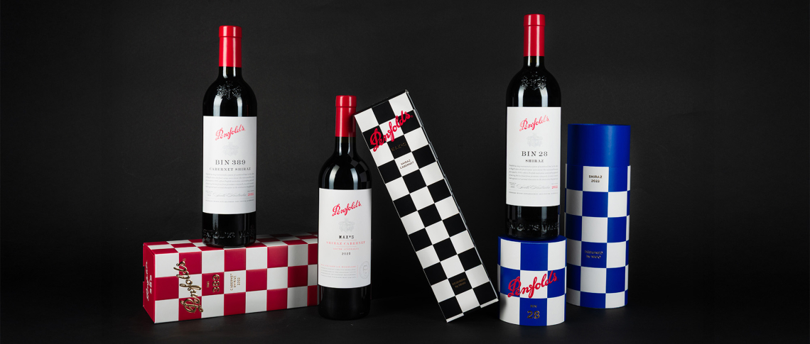 Drei Weinflaschen mit rot-weißem Etikett und gemusterten Verpackungen auf schwarzem Hintergrund.