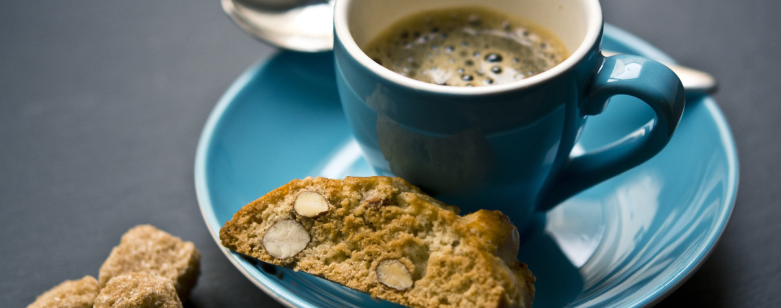 Blaue Tasse Kaffee mit Keks auf Unterteller und Zuckerwürfeln daneben.