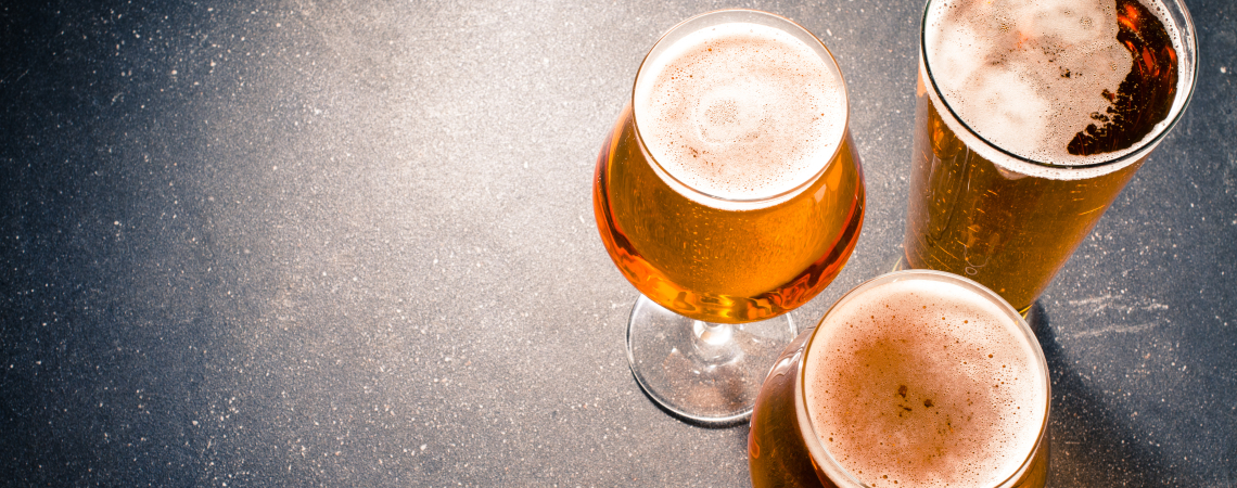 Drei Gläser Bier mit Schaumkrone auf dunklem, strukturiertem Hintergrund.