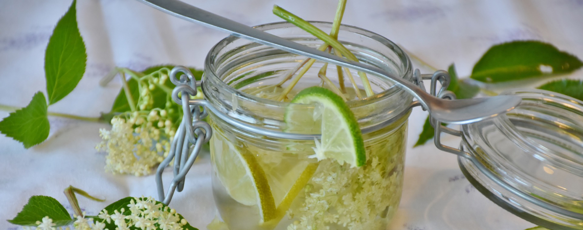 Glas mit Holunderblüten, Limettenscheiben und Blättern auf weißem Tischtuch.