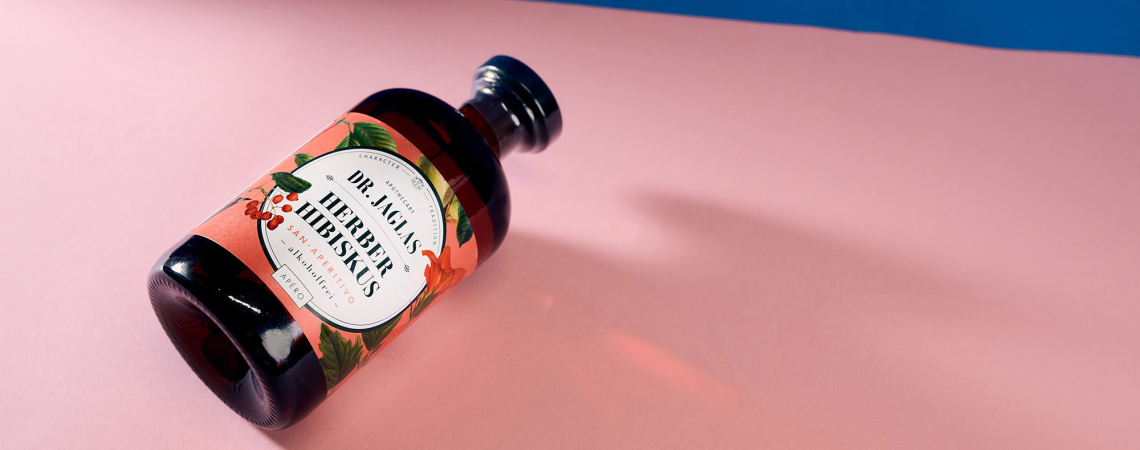 Braune Flasche mit floralem Etikett auf rosa und blauem Hintergrund.