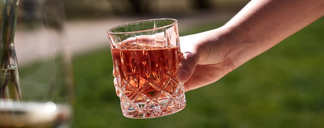 Hand hält ein Glas mit rosafarbenem Getränk, Tisch mit Snacks im Vordergrund.