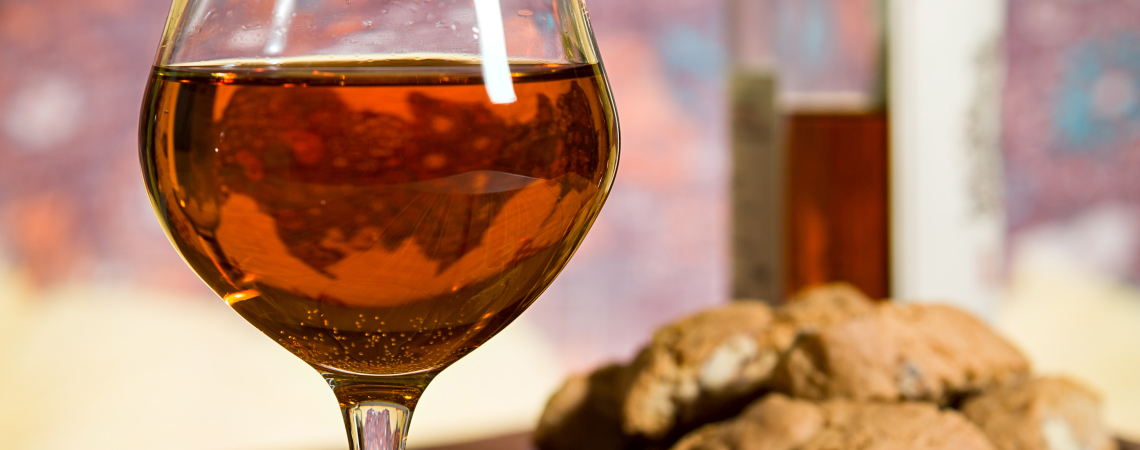 Weinglas mit goldenem Getränk, Weinflasche und Kekse auf Holztisch vor buntem Hintergrund.