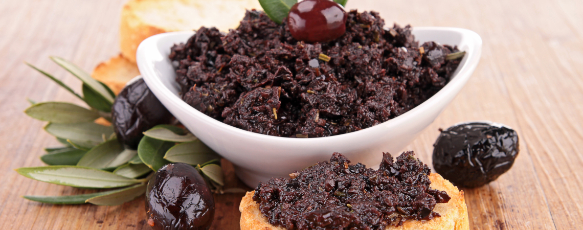 Weiße Schale mit dunklem Olivenaufstrich, garniert mit Oliven und knusprigem Brot.