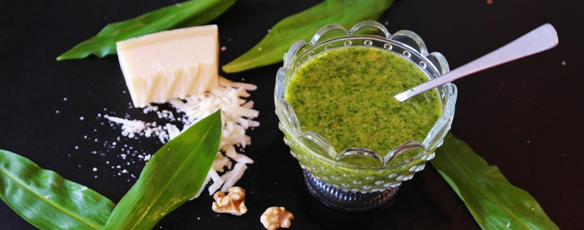 Grünes Pesto im Glas mit Parmesan, Bärlauchblättern und Walnüssen auf schwarzem Hintergrund.