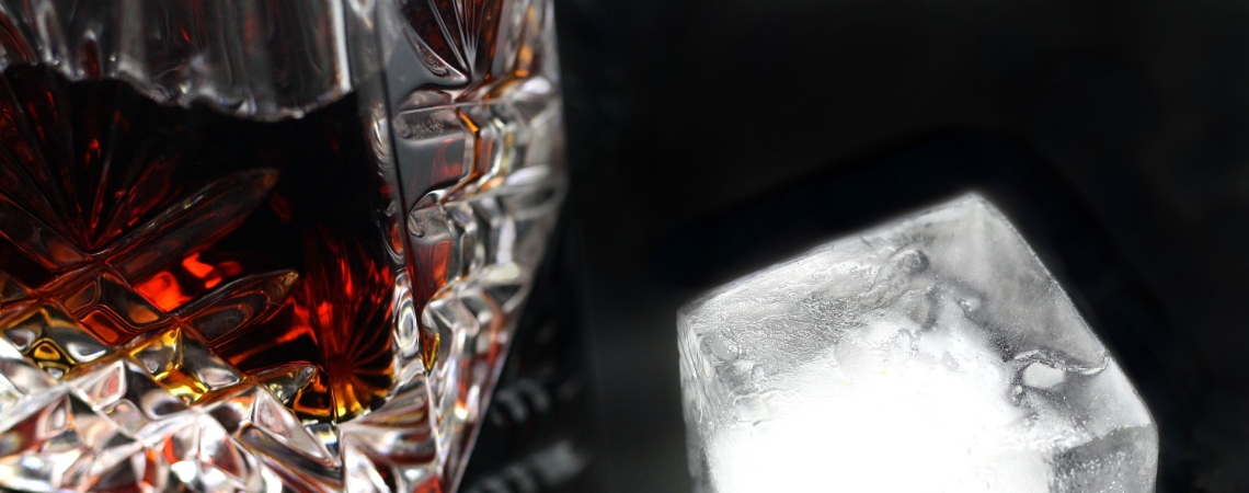 Kristallkaraffe mit bräunlicher Flüssigkeit und Eisklotz auf dunklem Untergrund.