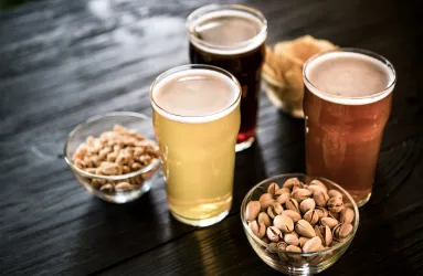 Drei Gläser Bier mit Nüssen und Chips auf dunklem Holztisch.