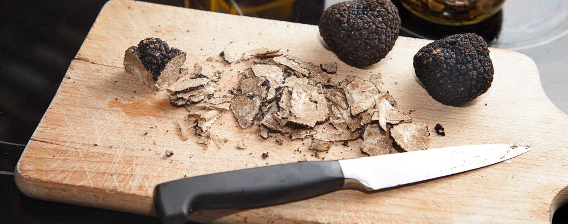 Schwarze Trüffel und ein Messer auf Holzbrett, neben Olivenöl und Glas.