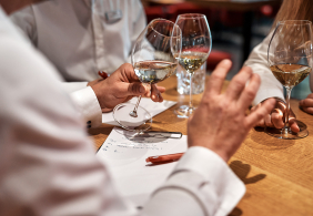 Personen in weißen Hemden diskutieren bei einem Glas Weißwein an einem Holztisch.