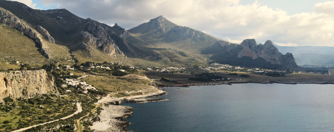 Küstenlandschaft mit felsigen Bergen, grüner Vegetation und blauem Meer unter bewölktem Himmel.