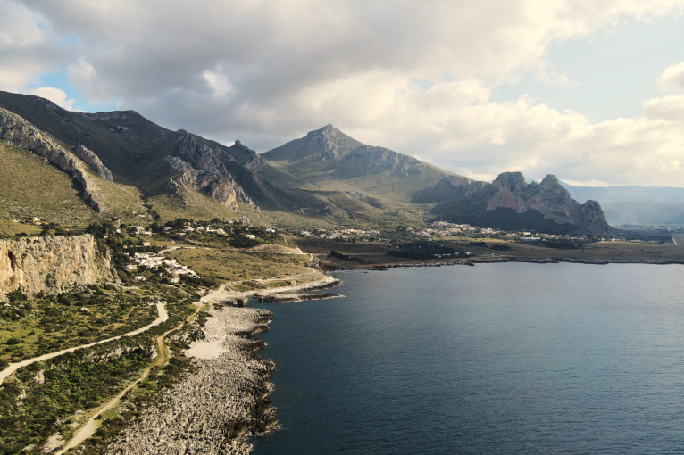 Küstenlandschaft mit felsigen Bergen, grüner Vegetation und blauem Meer unter bewölktem Himmel.