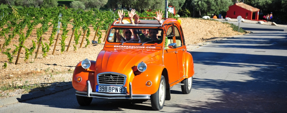 Leuchtend orangefarbener Oldtimer auf einer sonnigen Landstraße, Weinreben im Hintergrund.
