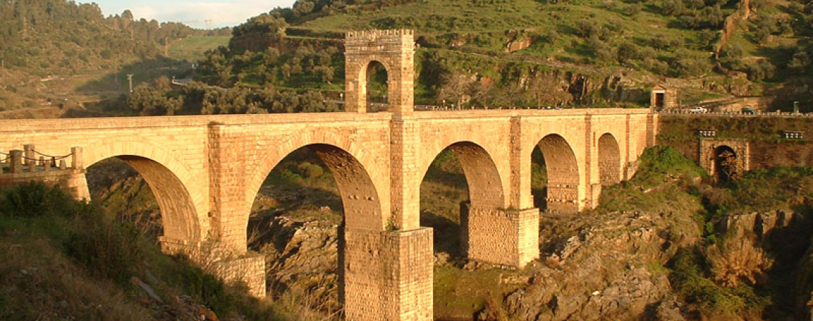 Steinbrücke über Fluss, umgeben von grünen Hügeln und antiken Ruinen.