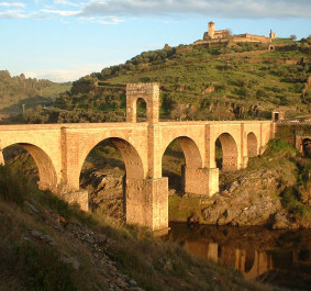 Steinbrücke über Fluss, umgeben von grünen Hügeln und antiken Ruinen.