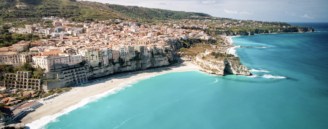 Küstenstadt mit pastellfarbenen Häusern und türkisblauer Meeresbucht unter blauem Himmel.