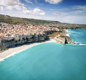 Küstenstadt mit pastellfarbenen Häusern und türkisblauer Meeresbucht unter blauem Himmel.