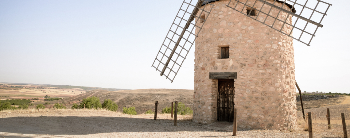 Steinwindmühle mit Metallflügeln in ländlicher Landschaft bei klarem Himmel.