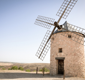 Steinwindmühle mit Metallflügeln in ländlicher Landschaft bei klarem Himmel.