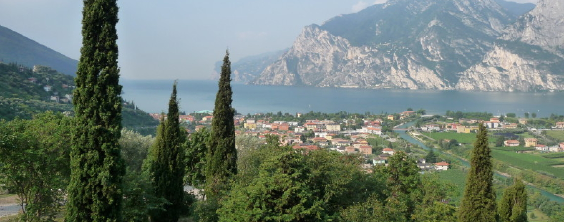 Blick auf grünes Tal, Zypressen, See und Berge bei klarem Himmel.