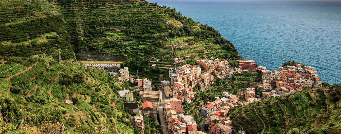 Bunte Küstenstadt mit terrassierten Feldern an einem grünen Hang, Meer im Hintergrund.
