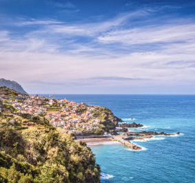 Küstenpanorama mit Dorf auf Hügeln, umgeben von blauem Meer und bewölktem Himmel.