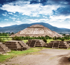 Weitläufige antike Pyramidenlandschaft mit grüner Vegetation und Bergen im Hintergrund.