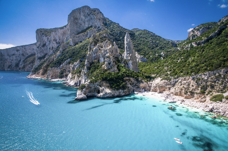 Klares türkisblaues Meer an felsiger Küste mit grünen Hügeln im Hintergrund.