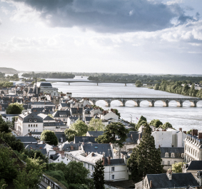 Stadtansicht mit Fluss, Brücke und Kirchturm unter bewölktem Himmel.