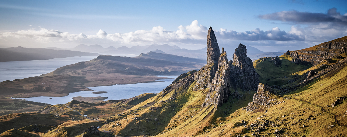 Felsformationen in grüner Hügellandschaft mit Blick auf einen See und Berge.