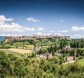 Hügelstadt mit historischer Architektur, umgeben von grünen Feldern und Zypressen unter blauem Himmel.