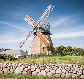 Historische Windmühle vor blauem Himmel, umgeben von Steinmauer und Grünfläche.