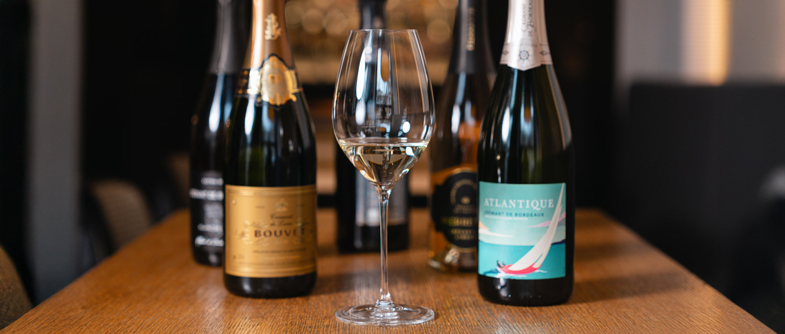 Fünf Sektflaschen und ein gefülltes Weinglas auf Holztisch im Hintergrund.