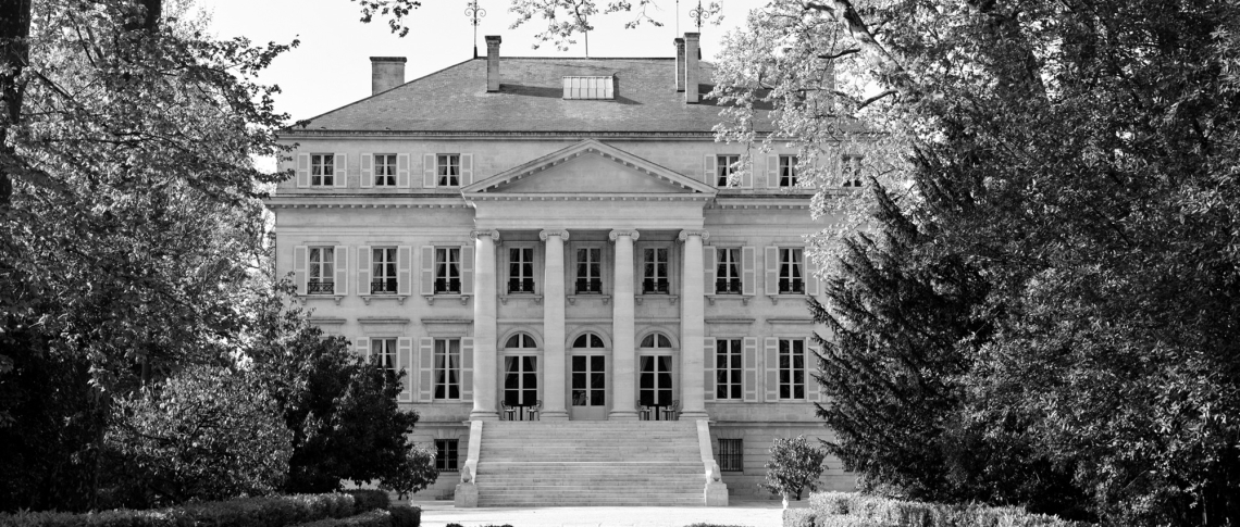 Historisches Herrenhaus mit Säulen, symmetrisch gestaltet, umgeben von Bäumen.