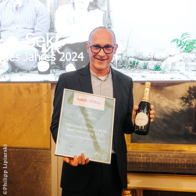 Mann in Anzug hält eine Urkunde und eine Flasche Sekt.