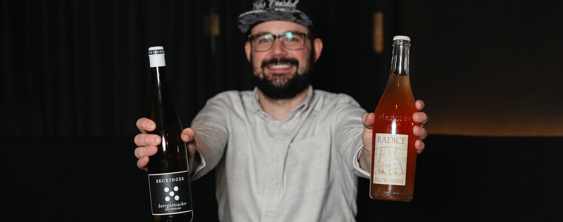 Mann in grauem Hemd hält zwei Weinflaschen vor dunklem Hintergrund.