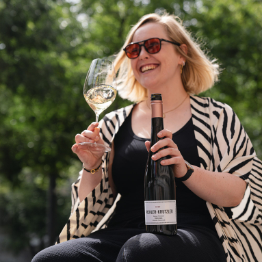 Frau mit Sonnenbrille hält Weinflasche und Weinglas in sonniger, grüner Umgebung.