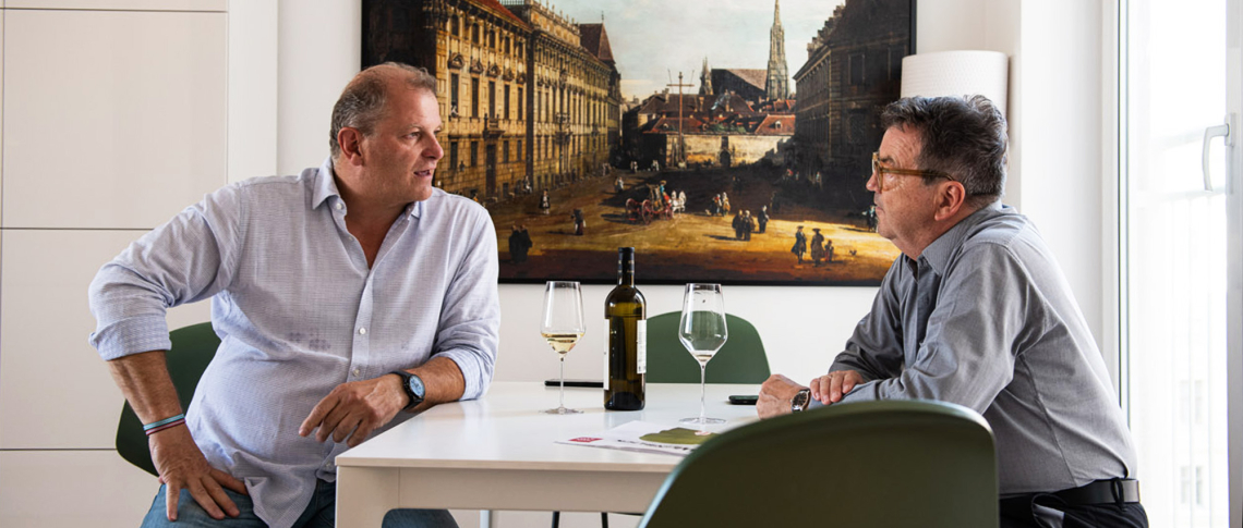 Zwei Männer unterhalten sich am Tisch mit Weinflaschen und Gemälde im Hintergrund.