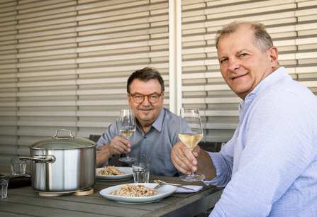 Zwei Männer genießen ein Mittagessen im Freien, Wein und Pasta auf dem Tisch.