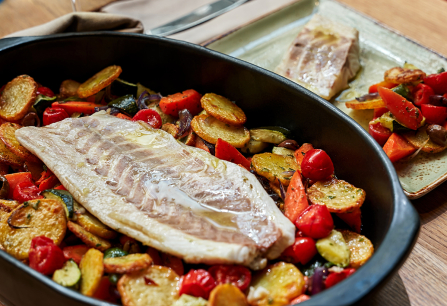 Gegrillter Fisch auf Ofengemüse in schwarzer Backform, daneben ein Glas Weißwein.