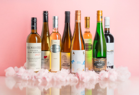 Sieben Weinflaschen auf rosafarbenem Hintergrund, umgeben von Wattewolken.