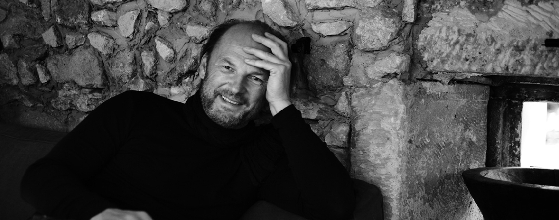 Schwarz-weiß Portrait eines lächelnden Mannes vor einer rustikalen Steinmauer sitzend.