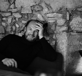Schwarz-weiß Portrait eines lächelnden Mannes vor einer rustikalen Steinmauer sitzend.