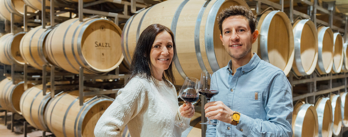 Zwei Personen stoßen in einem Weinkeller vor Eichenfässern mit Rotwein an.