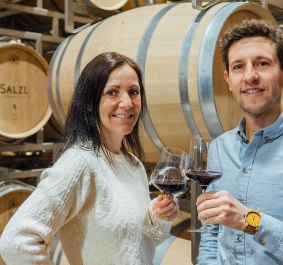 Zwei Personen stoßen in einem Weinkeller vor Eichenfässern mit Rotwein an.