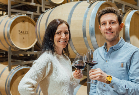 Zwei Personen stoßen in einem Weinkeller vor Eichenfässern mit Rotwein an.