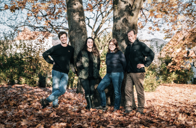Vier Personen stehen lächelnd vor einem herbstlichen Baum mit Laub am Boden.