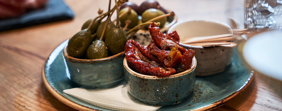 Rustikale Schälchen mit Kapern, getrockneten Tomaten und Oliven auf einem Holztisch.
