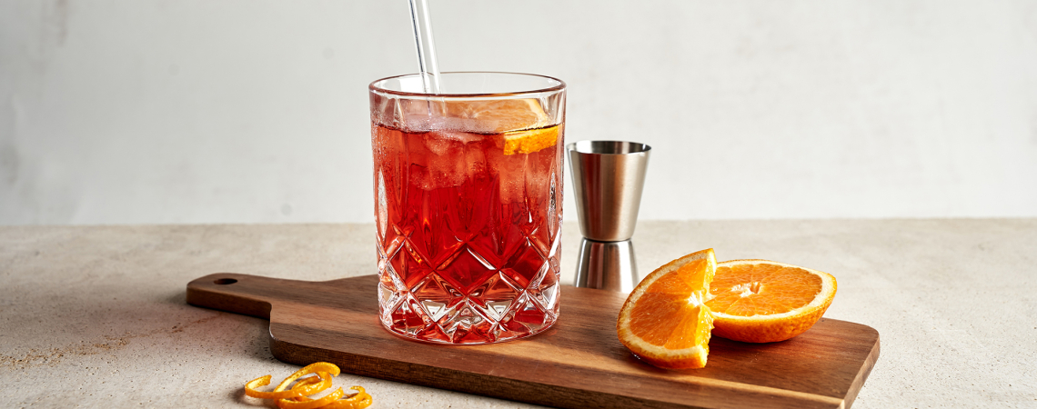 Roter Cocktail im Kristallglas auf Holzbrett mit Orangenscheiben.