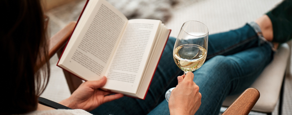 Person liest ein Buch und hält ein Glas Weißwein.